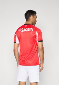 Rød fodboldtrøje med hvide accenter, sort kant og "SAGRES" trykt på ryggen. Bæres med hvide shorts. Let, åndbart stof.
