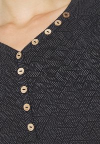 Schwarzes, gemustertes Stoffoberteil mit V-Ausschnitt und hölzernen Knöpfen vorne, das ein strukturiertes Design mit kleinen weißen Punkten hervorhebt.