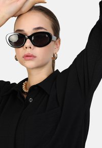 Zwarte cat-eye zonnebril met een glanzende afwerking en witte accenten. Draagster heeft een zwart shirt en een zware, ronde gouden ketting om.