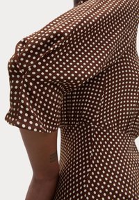 Abito marrone con pois bianchi, caratterizzato da maniche corte a sbuffo e vita arricciata, che mette in risalto un tessuto testurizzato.