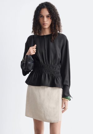 Femme aux cheveux bouclés portant un haut noir à manches longues avec une taille froncée, une jupe beige, tenant un sac à épaule noir et portant des bagues et des bracelets.
