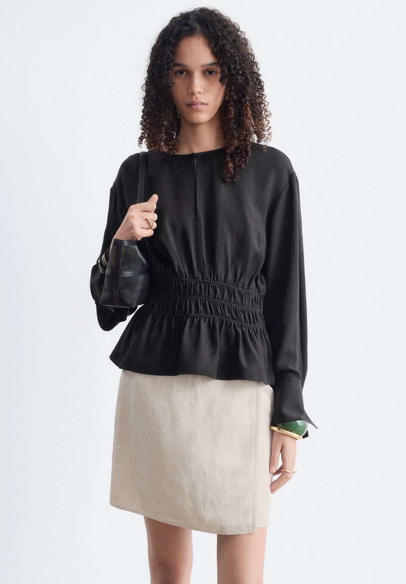 Femme aux cheveux bouclés portant un haut noir à manches longues avec une taille froncée, une jupe beige, tenant un sac à épaule noir et portant des bagues et des bracelets.