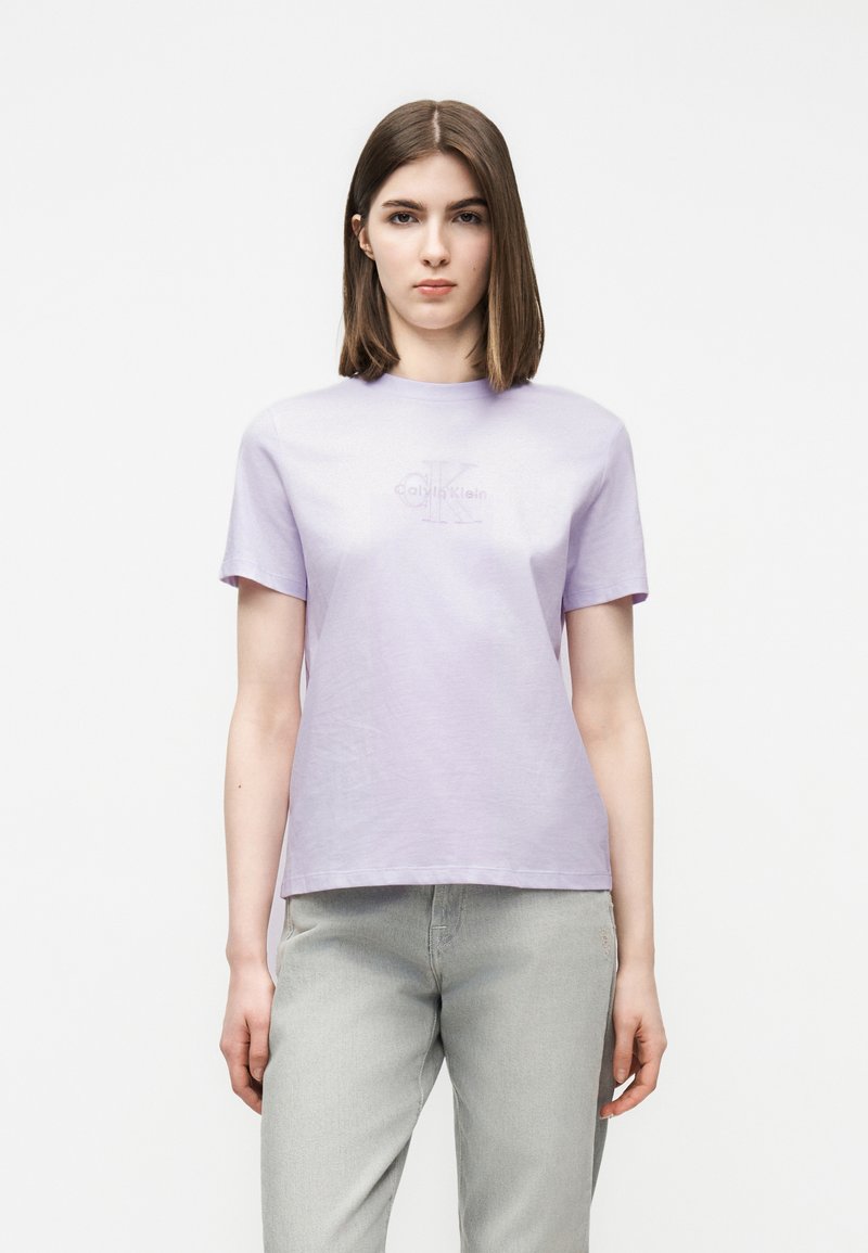 Γυναίκα με ίσια καστανά μαλλιά που φοράει ένα ανοιχτό μωβ T-shirt Calvin Klein και ανοιχτό γκρίζο παντελόνι, στέκεται μπροστά σε απλό λευκό φόντο.