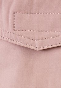 Ljusrosa tyg med en strukturerad yta, med en stickad ficka och en chevron-sömdetalj. Materialet verkar lätt och mjukt.