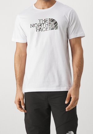 Mann trägt ein weißes Kurzarm-T-Shirt mit einem schwarzen gemusterten "The North Face"-Logo und schwarze Hose mit kleinem weißen Logo.