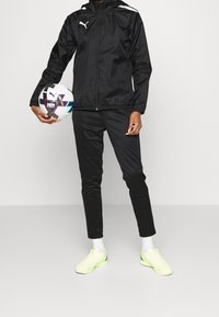Sort vindjakke med hætte og hvide detaljer, parret med sorte bukser og neongule sportssko, mens der holdes en fodbold.