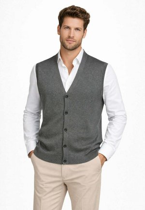 Uomo con gilet grigio senza maniche abbottonato sopra camicia bianca con pantaloni beige, mani nelle tasche, sfondo neutro.