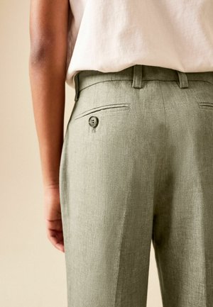 Close-up of person wearing light green linen trousers with a buttoned back pocket and a white tucked-in top.