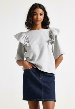 Nainen, joka pukeutunut vaaleanharmaaseen yläosaan, jossa on röyhelöiset olkapäät, ja tummaan siniseen farkkuhameeseen, seisoo yhdellä kädellä taskussaan.