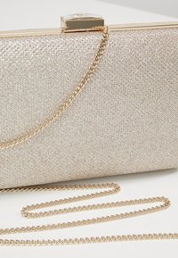 Rechteckige Clutch mit goldenem Glitzer, einem goldenen Kettenriemen und einem quadratischen Edelsteinverschluss oben.