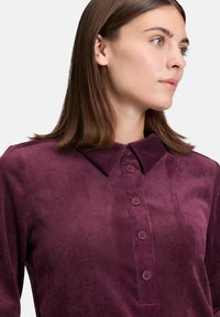 Junge Frau mit glattem braunem Haar, die ein lila Kordhemd mit Knöpfen trägt und nach rechts schaut, vor einem hellen Hintergrund.