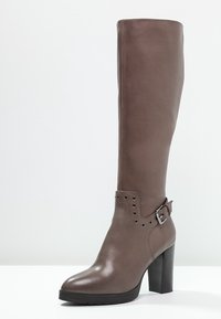 Bota de cuero marrón hasta la rodilla con un acabado pulido, acento de hebilla y tachuelas decorativas cerca del tobillo. Tacón grueso y alto.
