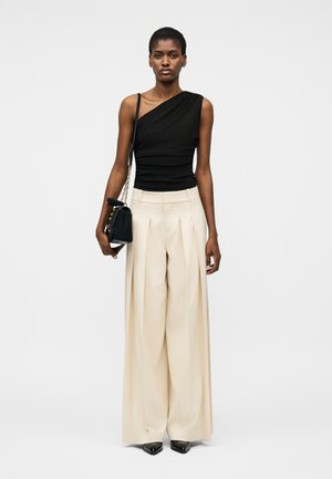 Ženska v črni enoramni majici, kremasto širokih plisiranih hlačah, črnih koničastih čevljih in s črno torbico z zlatim kovinskim trakom.