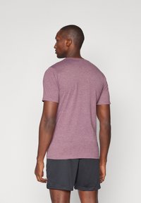 T-shirt court bordeaux à col rond, fabriqué en tissu doux et respirant ; porté avec un short de sport noir.
