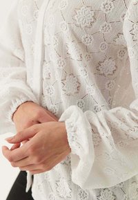 Biała haftowana bluzka z motywami kwiatowymi, teksturowaną tkaniną, elastycznymi mankietami i luźnym krojem, podkreślająca misternie wykonane koronki.