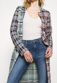 Abrigo de cuadros largo en tela multicolor que presenta azul, burdeos y verde, con un corte relajado y un diseño de abertura. Lleva puesto sobre una blusa beige y jeans.