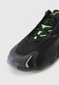 Gros plan d'une chaussure de sport noire avec dessus en mesh, accents vert néon, et lacets à motifs sur un fond clair uni.