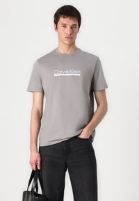 Camiseta de algodón gris con mangas cortas, que presenta un logo blanco de "Calvin Klein" en el pecho, combinada con jeans de mezclilla oscura y una bolsa negra.