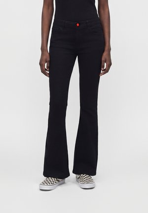 Persoană care poartă pantaloni evazați negri cu un nasture roșu, top negru și pantofi slip-on în șah alb-negru, stând pe un fundal simplu.