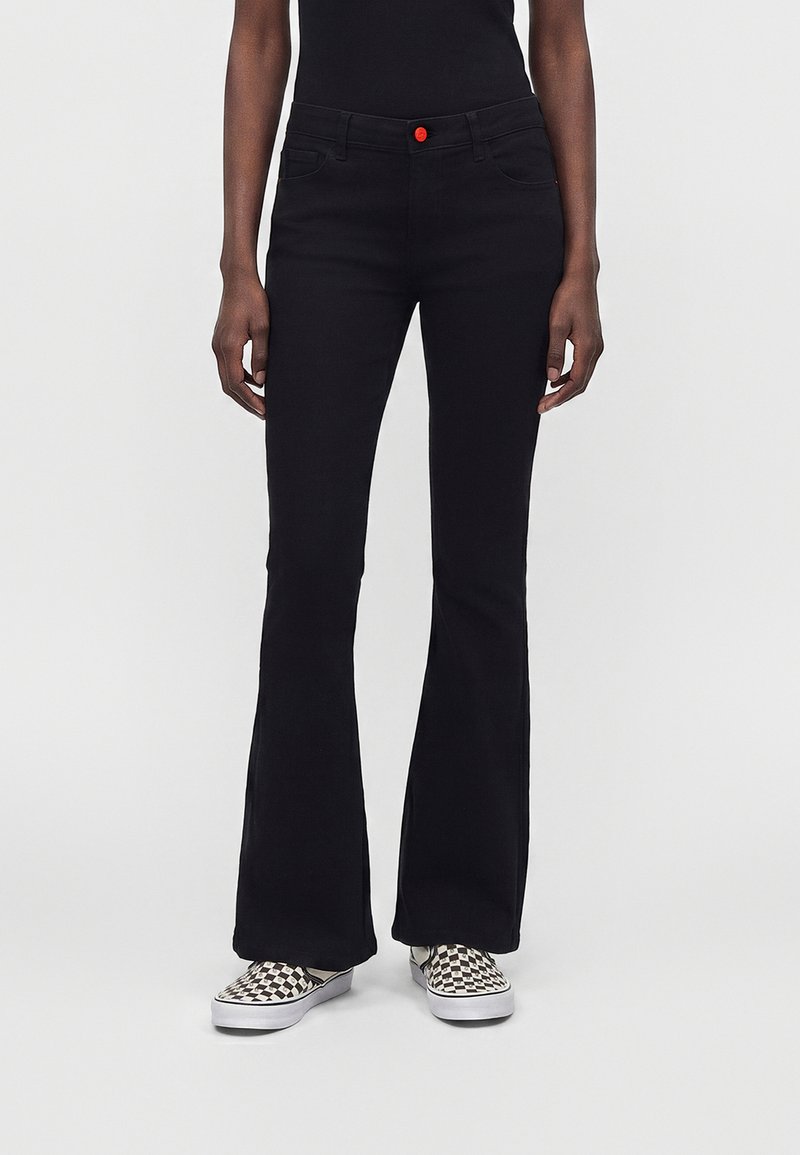 Person som har på seg svarte vide bukser med en rød knapp, svart topp og svart-hvite slip-on sko med sjakkbrettmønster, stående på ensfarget bakgrunn.