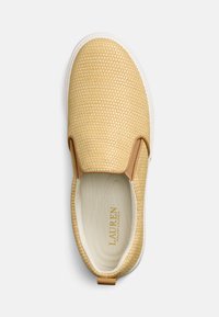 Beige vävd slip-on-sko med en tan läderdetalj vid hälen och vit gummisula; texturerad ovandel med varumärkeslogotyp inuti.