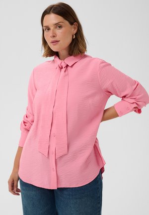 Woman with shoulder-length brown hair wearing a textured pink button-up blouse with a tied collar and blue jeans, standing with one hand on her hip.