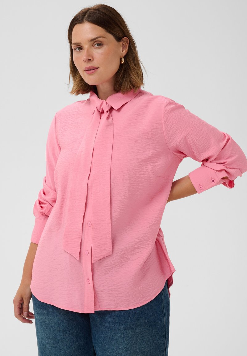 Femme aux cheveux bruns mi-longs portant un chemisier rose texturé à boutons avec un col noué et un jean bleu, debout avec une main sur la hanche.