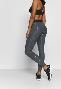 Szare legginsy z delikatnym, powtarzającym się wzorem "Nike", wysokim stanem i gładką teksturą, zestawione z czarnymi butami sportowymi.