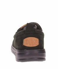 Zapato negro tipo slip-on con parte superior de ante, cuello acolchado y suela de goma. Presenta un parche de logo "Hey Dude" en color marrón claro en el talón.