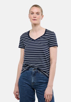 Femme portant un t-shirt à manches courtes à rayures bleu marine et blanches avec un col en V, rentré dans un jean bleu taille haute, debout devant un fond uni.