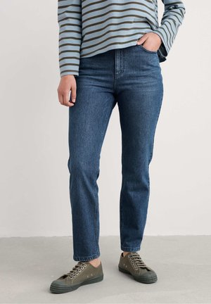 Person iført blå jeans, grønne lærreds sneakers med snøre og en langærmet stribet skjorte, står med den ene hånd i lommen.