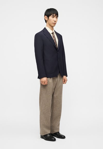 Άνδρας που φορά σκούρο σακάκι, ανοιχτό πουκάμισο, γραβατα με σχέδιο, φαρδιά μπεζ παντελόνια και μαύρα παπούτσια, στέκεται μπροστά σε απλό λευκό φόντο.