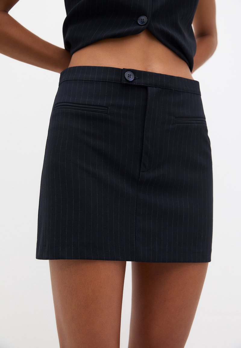 Mini-jupe noire à rayures avec une taille ajustée, présentant deux poches avant et une fermeture à boutons visible. Texture de tissu lisse.