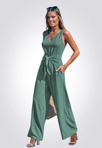 Mono verde con escote en V, detalle de lazo en la cintura, diseño de pierna ancha y tejido texturizado. Llevado con sandalias de tacón bloque blanco.