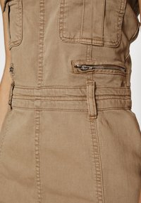 Tannfärgad jumpsuit med slim fit, synliga sömmar, dragkedjefickor och en strukturerad midjedesign. Bomullstyg med mjuk textur.