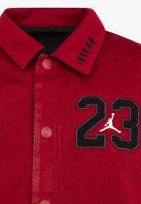 Rød mesh-trøye med svart "23" og Jumpman-logo. Har krage, knappelukking og embossed "JORDAN" på kragen.