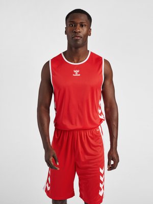Athletischer Mann, der ein rotes ärmelloses Sportoberteil und passende Shorts mit weißen Chevron-Designs an den Seiten trägt, steht vor einem einfarbigen Hintergrund.