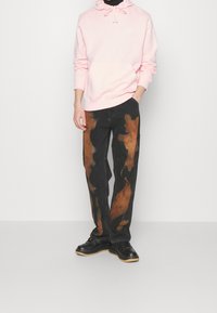 Ljust rosa hoodie med ficka framtill, kombinerad med svarta jeans med bruna abstrakta mönster, och svarta snörskor.