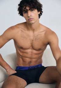 Caleçons boxers noirs avec une ceinture violette, arborant le logo Nike. Le modèle a des cheveux bouclés et un physique musclé et tonique.