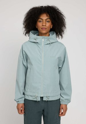 Woman with curly hair wearing a light blue zip-up hooded jacket with drawstrings and dark pants, standing against a plain background.