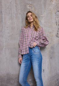 Jeune femme portant une chemise à carreaux rose et un jean bleu, debout contre un mur en béton brut, main dans la poche, l'air détendue.
