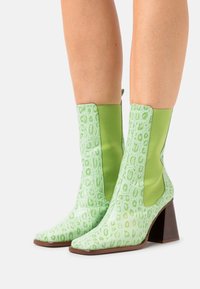 Botas de tobillo verdes con un patrón cocodrilo texturizado y brillante, paneles lisos en verde contrastante y un tacón cuadrado de color marrón medio. Diseño de punta cuadrada.