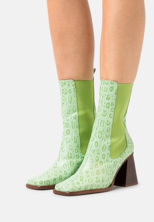 Botas de tobillo verdes con un patrón cocodrilo texturizado y brillante, paneles lisos en verde contrastante y un tacón cuadrado de color marrón medio. Diseño de punta cuadrada.