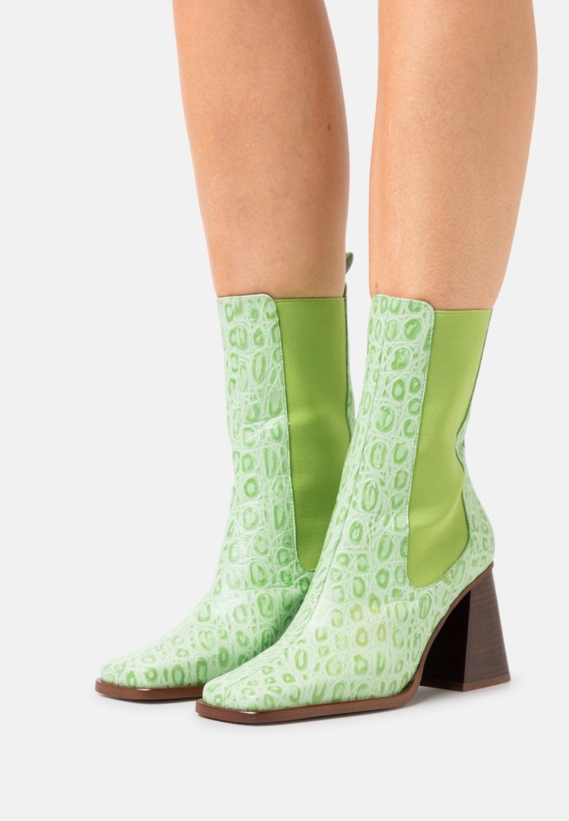 Botas de tobillo verdes con un patrón cocodrilo texturizado y brillante, paneles lisos en verde contrastante y un tacón cuadrado de color marrón medio. Diseño de punta cuadrada.