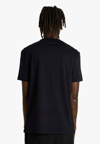 Persona con dreadlocks vista da dietro, che indossa una t-shirt nera a maniche corte semplice e pantaloni neri, con un tatuaggio visibile sul braccio sinistro.