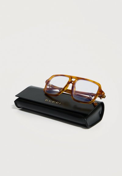 Ορθογώνια γυαλιά οράσεως με σχέδιο χελιδονόπτερου ξεκουράζονται σε μαύρη θήκη Gucci από δέρμα με ανάγλυφο χρυσό λογότυπο, σε λευκό φόντο.