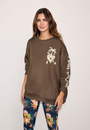 Kvinde iført en brun oversize sweatshirt med hvidt hjerte- og palmetrædesign, kombineret med farverige leggings med blomsterprint.