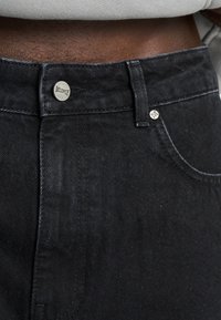 Jeans de mezclilla negra con cierre de botón metálico, marcados con "MILPLY" y un diseño clásico de cinco bolsillos con textura sutil.