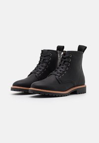 Bottines en cuir noir avec une finition texturée, présentant un laçage à l'avant, une fermeture éclair sur le côté et une semelle en caoutchouc marron contrastée.