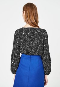 Blusa nera con motivi floreali e volti, maniche lunghe. Abbinata a una gonna blu a vita alta, con una texture liscia.